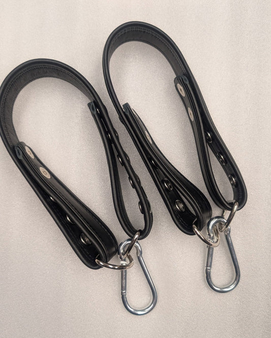 Two black leather straps with metal hooks on a beige surface