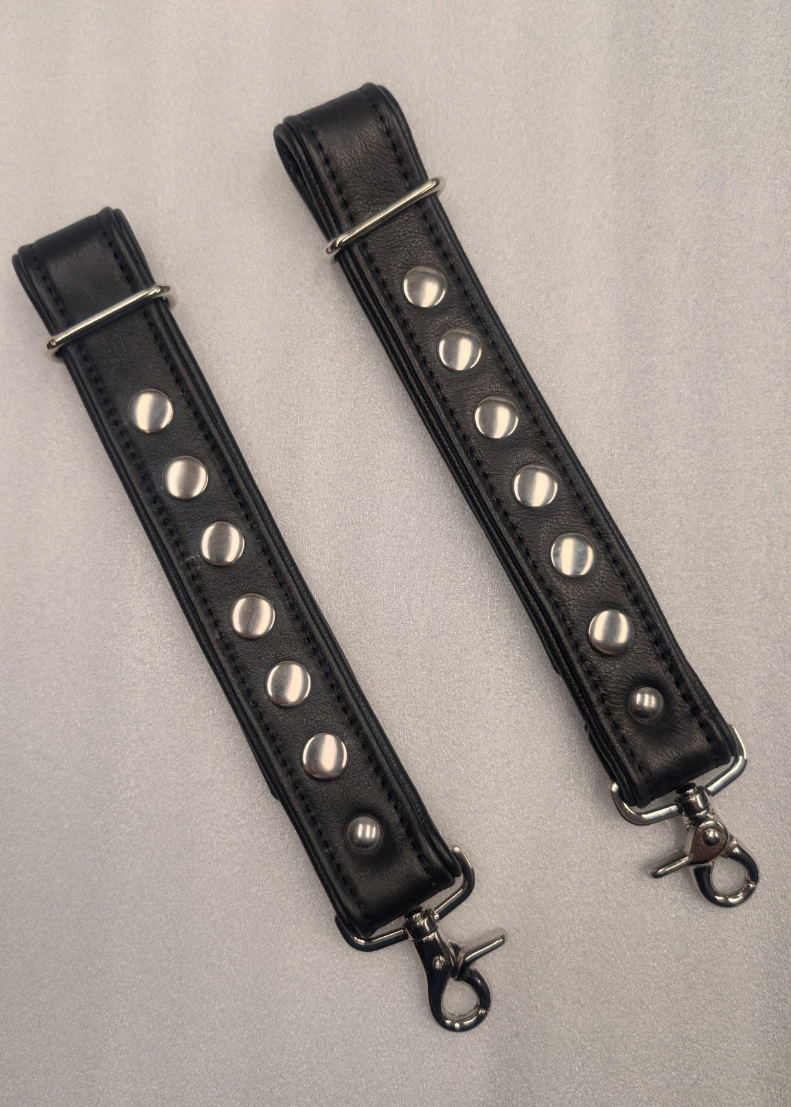 Two black leather straps with silver studs and metal clasps on a light gray background